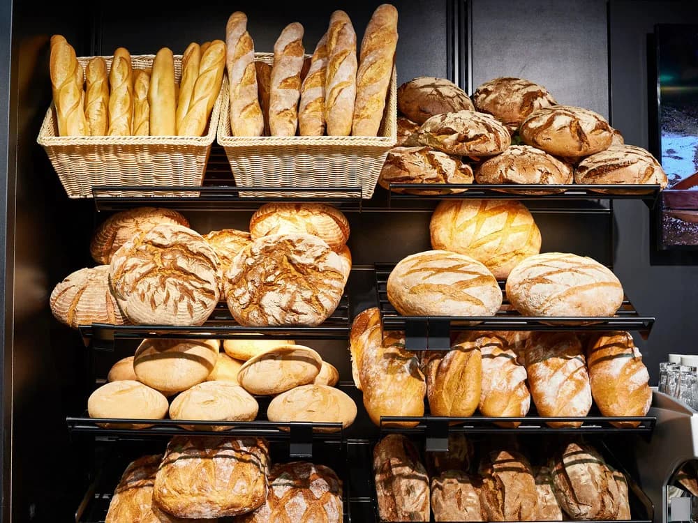Prateleiras escuras com cestos e bandejas cheias de diferentes tipos de pão numa padaria