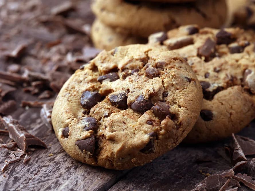 Cookies com pedaços de chocolate sobre superfície de madeira, cercados por lascas de chocolate
