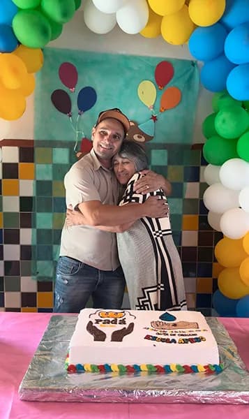 Um homem e uma mulher se abraçam sorrindo diante de um arco de balões coloridos, com um bolo decorado sobre a mesa