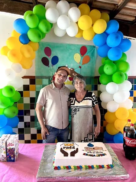 Um homem e uma mulher posam lado a lado sob um arco de balões coloridos, com um bolo decorado à frente e refrigerantes sobre a mesa