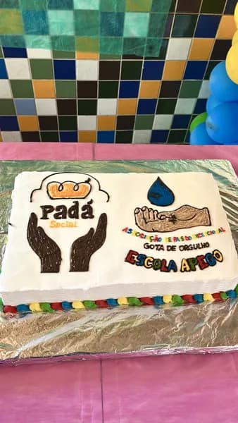 Bolo retangular decorado com o desenho de duas mãos, o logo do Padá Social e os dizeres Escola Apego, sobre papel alumínio