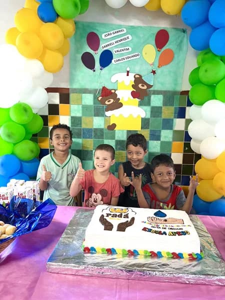 Quatro crianças sorridentes fazem sinal de positivo com o polegar atrás de um bolo decorado, diante de um cartaz com balões e nomes de aniversariantes