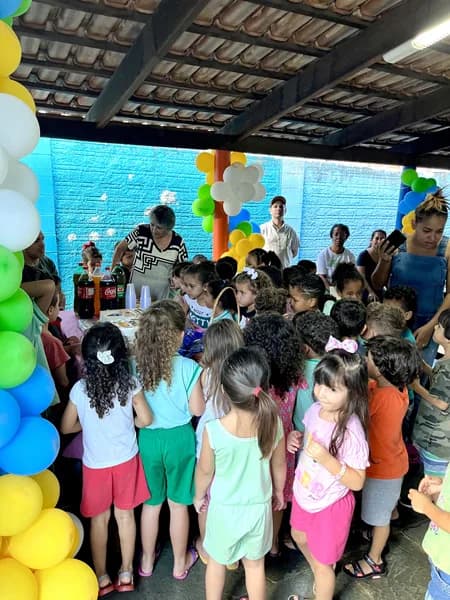 Grande grupo de crianças reunido ao redor de uma mesa com refrigerantes, sob arcos de balões, em pátio coberto da escola