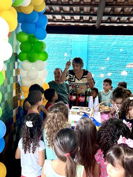 Duas pessoas adultas conversam com um grupo de crianças reunidas ao redor de uma mesa com refrigerantes e uma cesta de pães, sob decoração de balões
