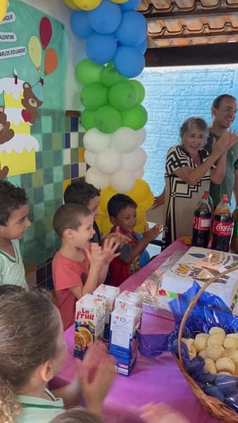 Grupo de crianças bate palmas diante de uma mesa com velas acesas, caixinhas de suco e uma cesta de pães, com adultos aplaudindo ao fundo