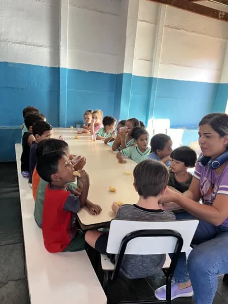 Crianças sentadas lado a lado em uma longa mesa comendo lanche, com uma pessoa adulta sentada na ponta da mesa