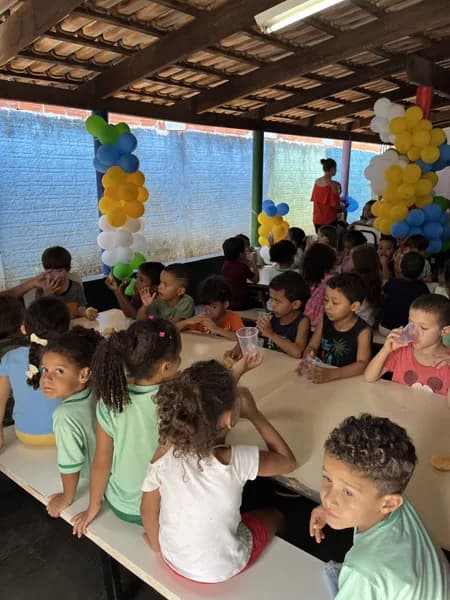Grupo de crianças sentado em mesas compridas em pátio coberto, com arranjos de balões ao fundo e adultos em pé observando