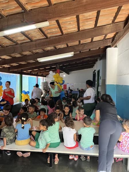 Crianças sentadas em fileiras em mesas compridas sob cobertura de madeira, com adultos em pé ao lado