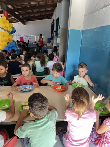 Crianças sentadas à mesa comendo com talheres em pratos coloridos, com outras crianças passando ao fundo
