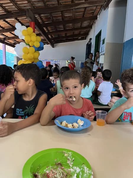 Um menino come de colher em um prato azul, sentado à mesa entre outras crianças, com adultos ao fundo
