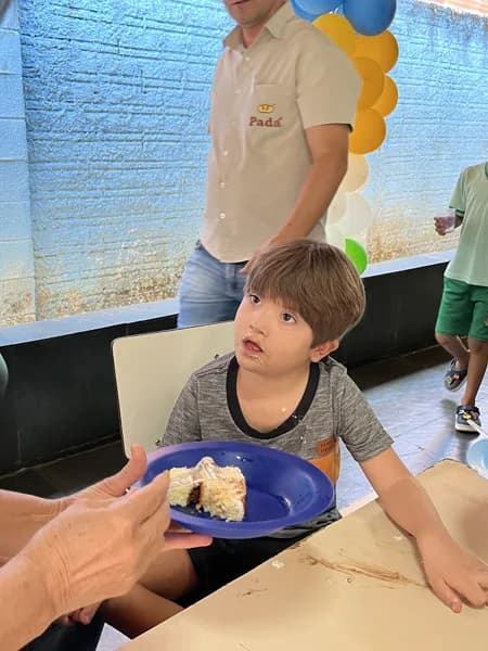 Um menino sentado à mesa olha para um prato de bolo estendido por uma mão adulta, com um homem de camisa Padá passando ao fundo