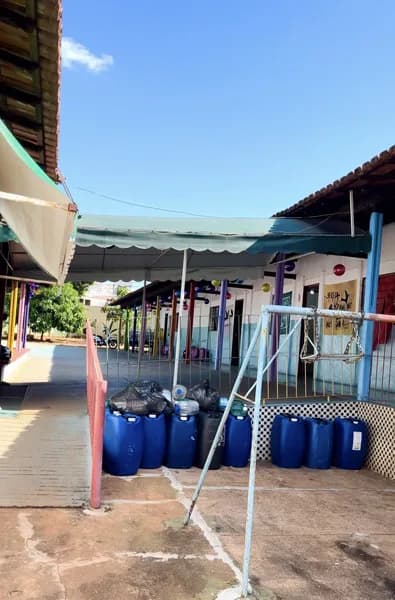 Pátio externo vazio da escola, com toldo verde, bombonas de água azuis enfileiradas e uma estrutura metálica com rede junto ao alambrado