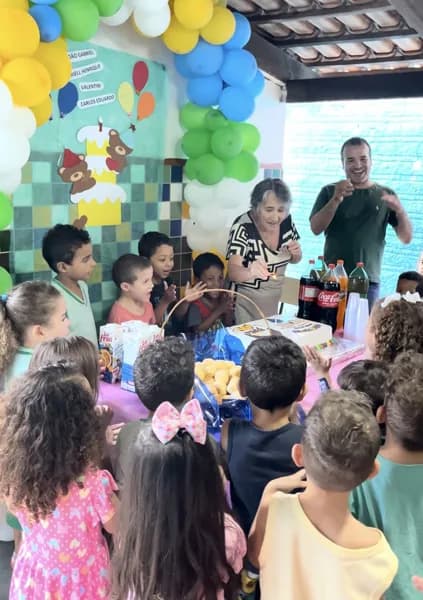 Grupo de crianças reunido ao redor de uma mesa com uma cesta de pães e refrigerantes, com um homem e uma mulher em pé atrás
